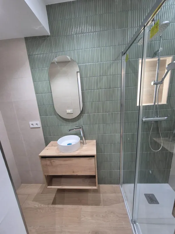 Baño reformado con azulejo verde, espejo ovalado y mueble de madera